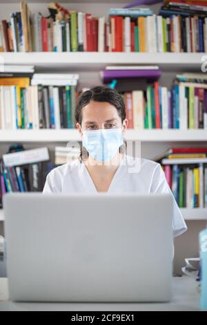 Ernst junge Ärztin trägt weiße Uniform und medizinische Maske Arbeiten auf Laptop in Latexhandschuhen sitzen am Schreibtisch in Moderne Klinik Stockfoto