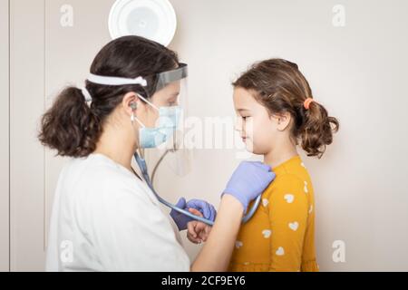 Seitenansicht der Ärztin in weißer Uniform und in Latexhandschuhe und Schutzmaske, die mit Stethoskop Lunge untersucht Kleines Mädchen Stockfoto