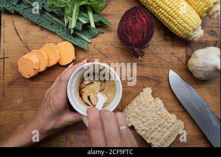Draufsicht einer Frau, die in einer Tasse veganes Dressing auf Holzschneidebrett mit verschiedenen Herbstgemüse und Tempeh-Protein macht. Stockfoto