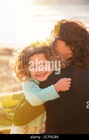 Seitenansicht der liebenden Frau hält niedlichen kleinen Mädchen mit Lockiges Haar beim Blick auf die Kamera im Freien Stockfoto