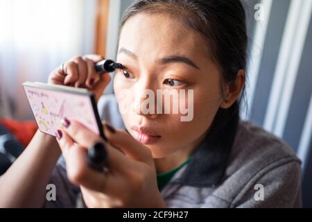 Junge asiatische Frau in Freizeitkleidung mit kleinen Spiegel in Hand Make-up und setzen schwarze Mascara auf Wimpern Stockfoto