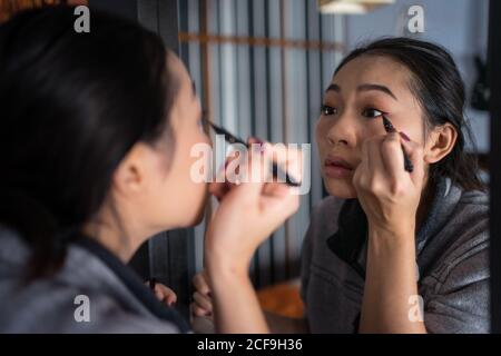 Moderne asiatische Frau in legere Kleidung Anwendung schwarzen Eyeliner während Zu Hause vor dem Spiegel stehen und sich vorbereiten Für die Arbeit Stockfoto