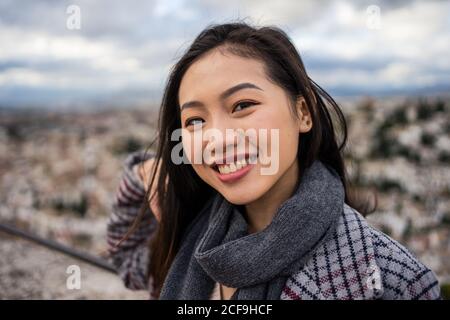 Happy Asian Dame in stilvollen Mantel und Schal lächelnd und Blick auf die Kamera, während Sie auf verschwommenem Hintergrund von Alter stehen Stadt am bewölkten Tag Stockfoto