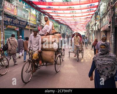 Bangladesch - Januar 20 2019: Traditionelle Rikschas mit ethnischen Männern und vollen Säcken, die auf Asphaltstraße durch Marktstraße in der Stadt reiten Stockfoto