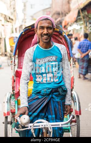 Bangladesch - Januar, 25 2019: Fröhlicher ethnischer Mann in lässiger Kleidung lächelnd und mit Blick auf die Kamera, während er auf der Straße der Stadt die traditionelle Rikscha fährt Stockfoto