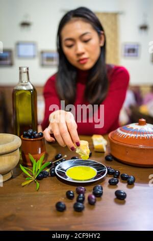 Nachdenkliche junge asiatische Frau sitzt und taucht Brot in Olivenöl in Teller auf dem Tisch mit Gerichten und Oliven Stockfoto