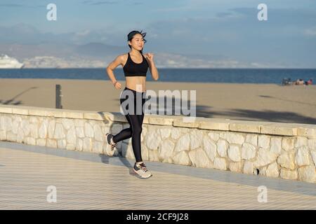 Motivierte junge Sportlerin in aktiver schwarzer Kleidung und Sneakers Jogging entlang felsigen Zaun am Strand an der leeren Küste Stockfoto