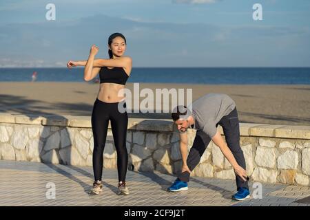 Zufrieden junge sportliche Paar stehend Stretching in Sportbekleidung mit Blaues Meer und Himmel auf verschwommenem Hintergrund Stockfoto