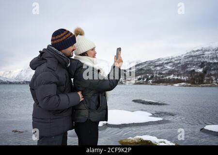 Junges Paar, Frau und Mann in warmen Kleidern, die mit dem Smartphone von der atemberaubenden winterlichen Landschaft des Hochlandes geschossen wurden, während sie am Ufer des Kaldfjords-Sees in Norwegen standen Stockfoto