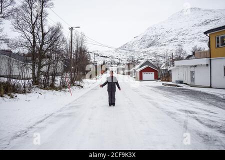 Alter Mann steht allein auf leeren rutschigen Straße gegen kleine Rote Landhäuser und grauer Himmel auf Ersfjordbotn See in Winter Stockfoto