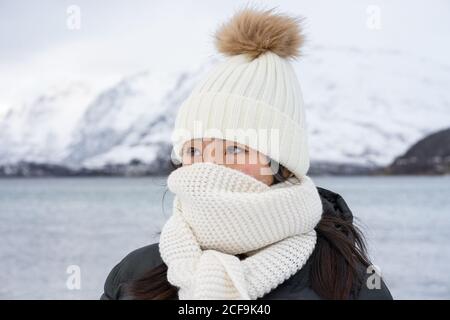 Junge Frau mit schwarzer Jacke und weißem warmen Hut und Schal, die vor grauem, unscharfem Hintergrund in Norwegen auf die Kamera schaut Stockfoto