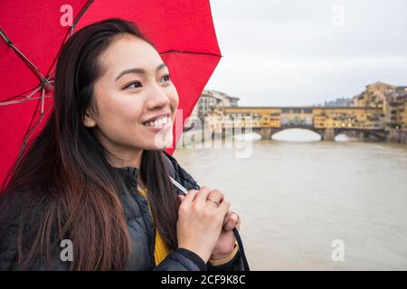Glückliche ethnische Frau mit rotem Schirm lächelnd und wegblickend, während sie an bewölktem Tag in Florenz, Italien, auf der Brücke gegen den Fluss und die Ponte Vecchio stand Stockfoto