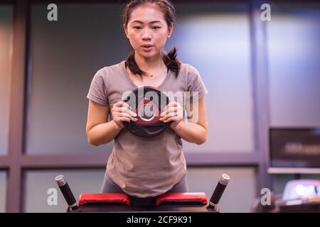 Starke junge ethnische Sportlerin in aktiver Kleidung, die sich auf der Bank lehnt und beim Training im modernen Fitnessstudio Hyperextension mit Hantelscheibe macht Stockfoto