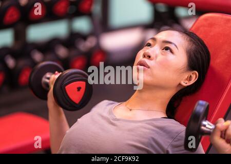 Leistungsstarke ethnische Athletin in Sportbekleidung tun Hanteln drücken auf Schrägbank mit Blick auf das moderne Fitnesscenter Stockfoto