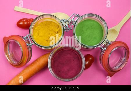 Draufsicht auf isolierte Einmachgläser und Holzlöffel mit gelbem Kurkuma, roter Rote Bete und grünem Spinatpulver, rosa Hintergrund. Stockfoto