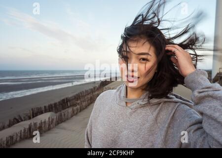 Calm junge asiatische Frau in Casual tragen berühren Haare und Blick auf die Kamera, während Sie in der Nähe der Küste unter bewölktem Himmel stehen An windigen Tagen Stockfoto