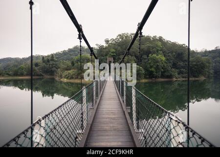 Hängebrücke mit Holzboden und Metallgitter über groß see und weitläufige grüne Wälder unter weißem Himmel am Nachmittag Stockfoto