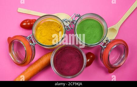 Draufsicht auf isolierte Einmachgläser und Holzlöffel mit gelbem Kurkuma, roter Rote Bete und grünem Spinatpulver, rosa Hintergrund. Stockfoto