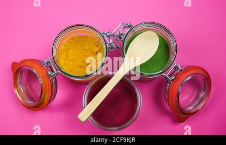 Draufsicht auf isolierte Einmachgläser und Holzlöffel mit gelbem Kurkuma, roter Rote Bete und grünem Spinatpulver, rosa Hintergrund. Stockfoto