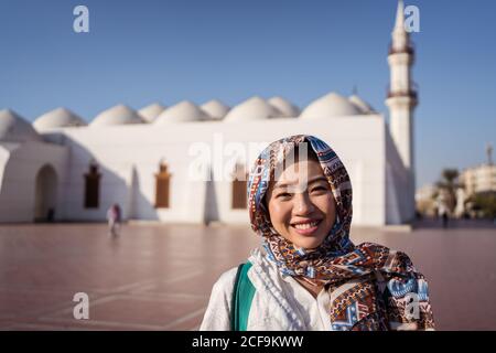 Glückliche junge asiatische Frau in traditionellen Kopftuch lächelnd und suchen Bei der Kamera, während auf dem Platz gegen Islam Moschee in stehen Jeddah Stadt in Saudi-Arabien Stockfoto
