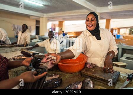 Jinja, Uganda - 1. Oktober: Hilfreiche afroamerikanische Frau, die großen Fisch hält und dem Käufer auf dem Marktplatz zeigt Stockfoto