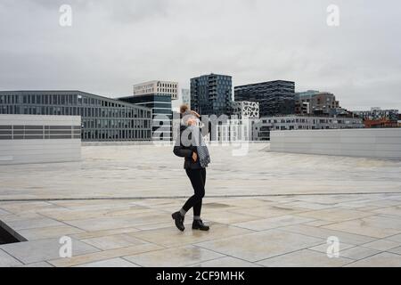 Fröhliche junge ethnische Frau, die Winterkleidung trägt und auf dem platz am Opernhaus von Oslo steht Stockfoto