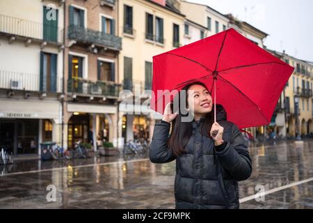 Junge asiatische weibliche Reisende in warmer Kleidung Sightseeing mit roten Regenschirm mit alten Gebäuden auf verschwommenem Hintergrund bei Padova at Italien Stockfoto