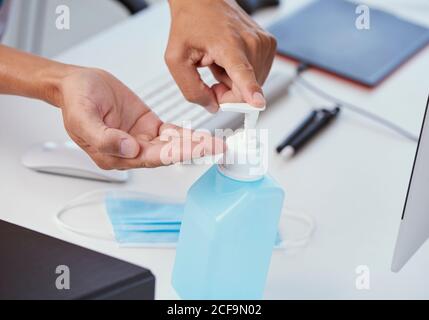 Nahaufnahme eines jungen kaukasischen Mannes an seinem Schreibtisch Seine Hände mit einem Händedesinfektionsmittel aus einer blauen Flasche desinfizieren Stockfoto