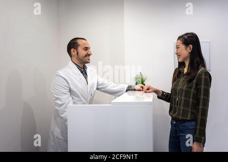 Seitenansicht der positiven männlichen Arzt in weißen Kleidern stehen An der Theke in der Klinik und im Gespräch mit lächelnden asiatischen Frau Patient Stockfoto