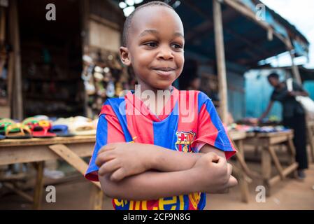 Ruanda, Afrika - 14. Dezember 2019: Von unten ein friedlicher afrikanischer Junge mit gekreuzten Armen im Fußballspieler-T-Shirt, der auf der lokalen Marktstraße steht und wegschaut Stockfoto