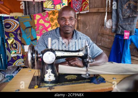 Ruanda, Afrika - 14. Dezember 2019: Gutherzigen afroamerikanischen Mann in Freizeitkleidung Nähkleidung auf Maschine in lokalen Markt Blick auf Kamera Stockfoto