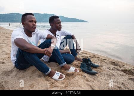 Ruanda, Afrika - 14. Dezember 2019: Junge afroamerikanische Männer in lässiger Kleidung sitzen zusammen am Sandstrand und blicken tief in Gedanken davon Stockfoto