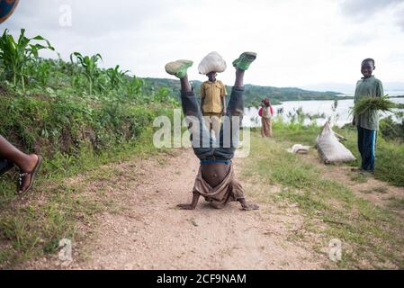 Ruanda, Afrika - 14. Dezember 2019: Afrikanischer Junge in Casual Wear Pause Tanz auf Händen stehen während der Arbeit in Plantage mit anderen Kindern Stockfoto