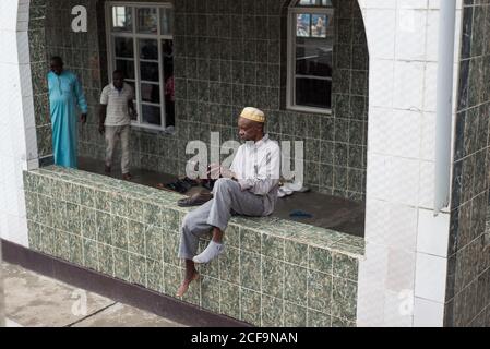 Ruanda, Afrika - 14. Dezember 2019: Von oben der religiöse afroamerikanische Mann in lässiger Kleidung tragen Schuhe sitzen auf dem Zaun der Moschee nach dem Gebet Stockfoto