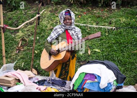 Ruanda, Afrika - 14. Dezember 2019: Ruhige afroamerikanische Frau in farbenfrohen einfachen Kleidern, die Gitarre spielen und Kleidung im Marktstand verkaufen und dabei die Kamera betrachten Stockfoto