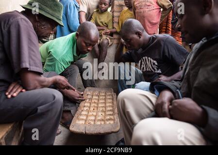 Ruanda, Afrika - 14. Dezember 2019: Gruppe von fokussierten afrikanischen Männern spielen Brettspiel auf der Straße im Dorf sitzen Stockfoto