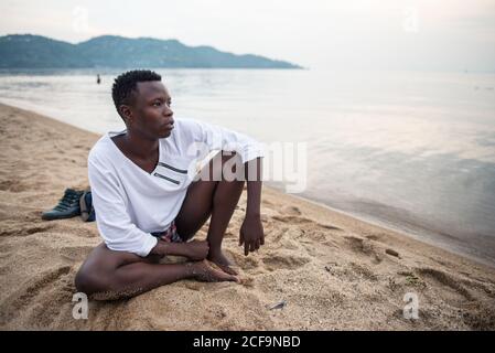 Ruanda, Afrika - 14. Dezember 2019: Afroamerikanischer Typ in Casual Wear, der am Sandstrand sitzt und nachdenklich davonschaut Stockfoto