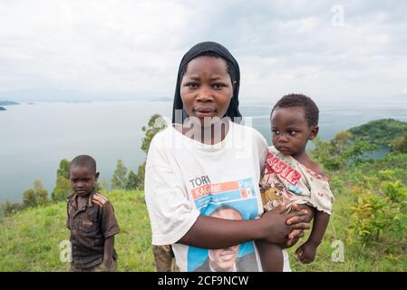 Ruanda, Afrika - 14. Dezember 2019: Ernste afroamerikanische Frau in einfacher Kleidung mit kleinem Kind an den Händen stehend mit Kind an der grünen Küste und Blick auf die Kamera Stockfoto