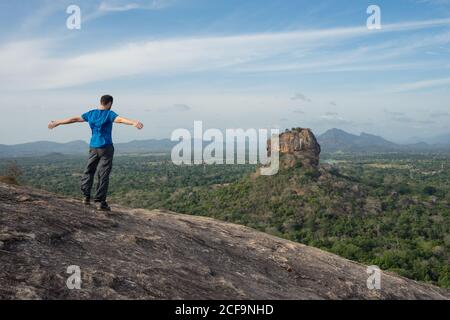 Rückansicht des unkenntlichen Mannes, der die Arme ausstreckt und eine fantastische Aussicht genießt Der einsamen Felsenfestung Sigiriya in der Mitte des Plateaus mit Dichter grüner tropischer Wald vom Berg Pidurangala unter blauem Himmel In Sri Lanka Stockfoto