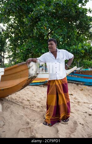 Tangalle, Sri Lanka - 28. Juli 2019: Ältere ethnische Fischer in lässiger leichter Kleidung, die auf dem Boot lehnt und wegschaut Stockfoto