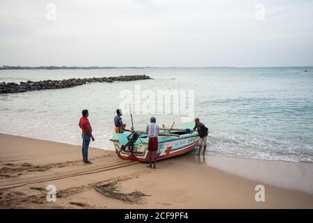 Tangalle, Sri Lanka - 28. Juli 2019: Ethnische Fischerleute in legerer Kleidung, die blaues Boot an die sandige, leere Küste treiben Stockfoto