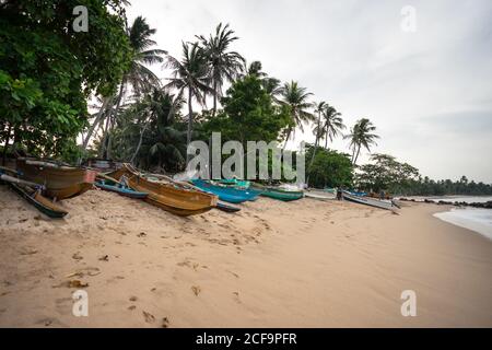 Tangalle, Sri Lanka - 28. Juli 2019: Leerer Sandstrand mit bunten Holzbooten und grünen exotischen Pflanzen bei bewölktem Wetter Stockfoto