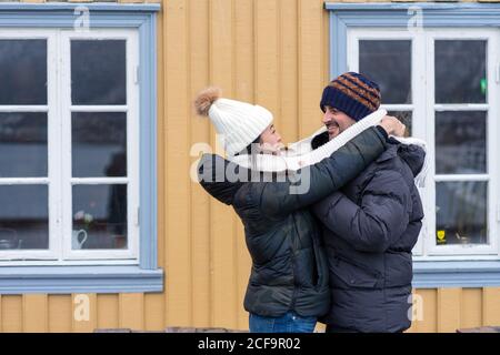 Seitenansicht des multiethnischen fröhlichen Paares in warmer Kleidung stehend Und Umarmung mit ländlichem Haus auf Hintergrund bei Tromso in Norwegen Stockfoto