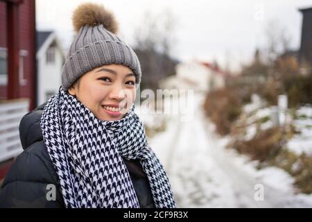 Fröhliche asiatische weibliche Reisende in warmer Kleidung lächelnd und suchen Bei der Kamera mit Berg auf verschwommenem Hintergrund bei Tromso in Norwegen Stockfoto