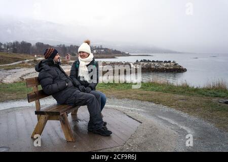 Gefrorene junge Freunde in warmen Kleidern sitzen auf einer Holzbank an der Hochküste und blicken in die Ferne des Meeres bei bewölktem Wetter in Tromso, Norwegen Stockfoto