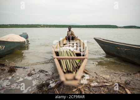 Uganda - November, 26 2016: Afrikanischer Junge in schmutziger Casual-Kleidung sitzt in schäbigen Boot mit Holzstöcken in der Nähe der Küste des Flusses Stockfoto