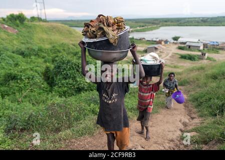 Uganda - November, 26 2016: Afrikanischer Junge mit Becken auf dem Kopf und Blick auf die Kamera während der Plantage in der Nähe von schwarzen Freunden Stockfoto