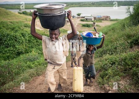 Uganda - November, 26 2016: Afrikanischer Junge mit Becken auf dem Kopf und Blick auf die Kamera während der Plantage in der Nähe von schwarzen Freunden Stockfoto