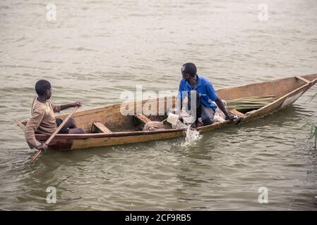 Uganda - November, 26 2016: Männlicher Teenager rudert, während erwachsener Mann Wasser aus dem alten Boot ausbackt und während der Fahrt auf dem schmutzigen Fluss wegschaut Stockfoto