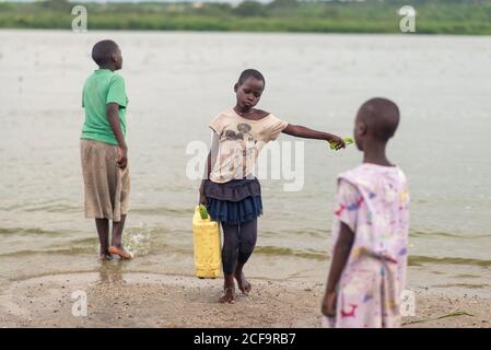 Uganda - November, 26 2016: Kleines afrikanisches Mädchen, das schweren Kanister mit Wasser aus dem Fluss trägt und an der nassen Küste in der Nähe von Mitkindern im Dorf läuft Stockfoto